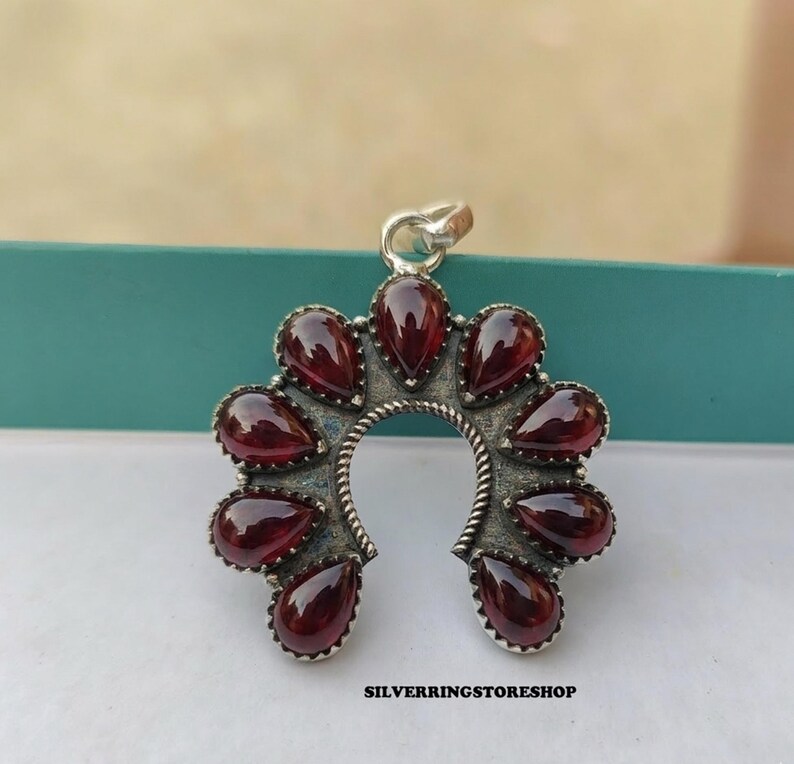 Pu&ograve; includere: Un ciondolo in argento con un design a ferro di cavallo, adornato con pietre preziose rosse a forma di goccia. Il ciondolo ha un bordo argentato strutturato e un piccolo anello per una catena. Il testo "SILVERRINGSTORESHOP" &egrave; in basso.