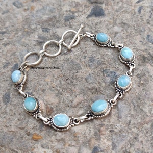 May include: A silver bracelet featuring oval, light blue gemstones. The gemstones are set in a decorative silver frame and linked together with silver chain. The bracelet has a toggle clasp. The text "silverringstore.shop" is visible.