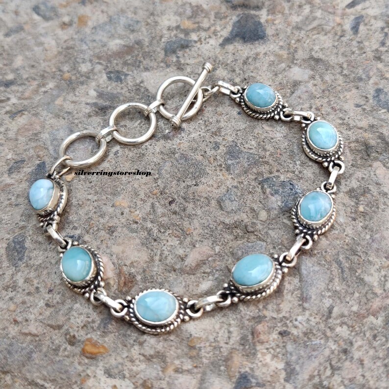 May include: A silver bracelet featuring oval, light blue gemstones. The gemstones are set in a decorative silver frame and linked together with silver chain. The bracelet has a toggle clasp. The text "silverringstore.shop" is visible.