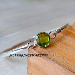 May include: A silver bangle bracelet with a large, round, faceted, olive-green gemstone. The bracelet has a simple, elegant design with a polished finish. The text "SILVERRINGSTORESHOP" is printed on the wooden surface.