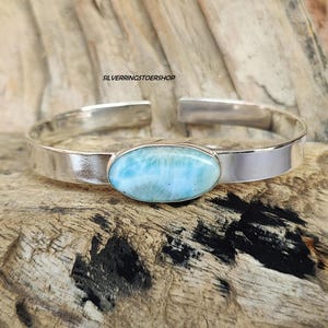 Pode incluir: Um bracelete de prata com uma pedra oval de cor azul claro. A pedra tem um padrão de redemoinho e está engastada em um bezel de prata.