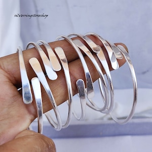 May include: A set of six silver bangle bracelets, each with a unique, slightly curved, flat design. The bracelets are stacked on top of each other, creating a layered look.