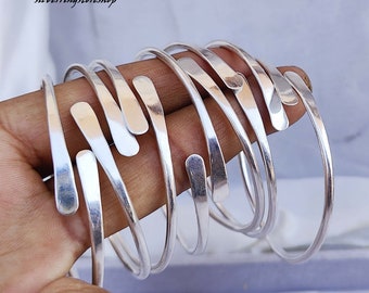 Puede incluir: Un conjunto de seis pulseras de plata, cada una con un diseño plano ligeramente curvado único. Las pulseras están apiladas una encima de la otra, creando un aspecto en capas.
