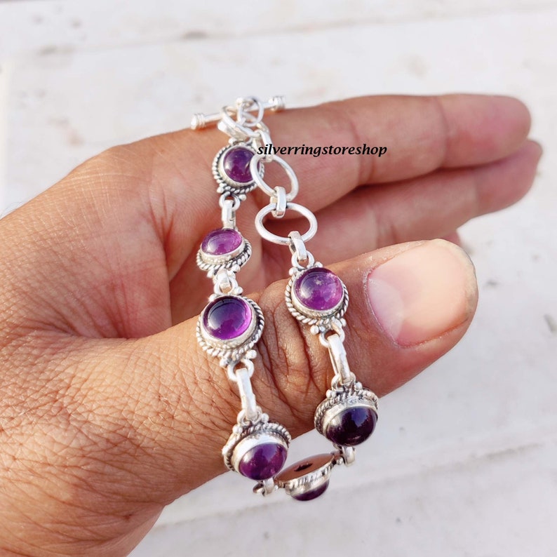 May include: A silver bracelet featuring multiple round, deep purple gemstones. The bracelet has a toggle clasp and is held in a hand. The gemstones are set in ornate silver settings, creating a vintage look.