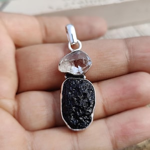 Puede incluir: Un colgante de plata con un cristal de cuarzo transparente sobre una roca volc&aacute;nica negra.
