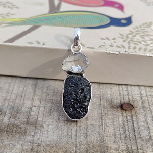 Puede incluir: Un colgante de plata con un cristal de cuarzo transparente y una piedra de obsidiana negra. El colgante est&aacute; sobre una superficie de madera.