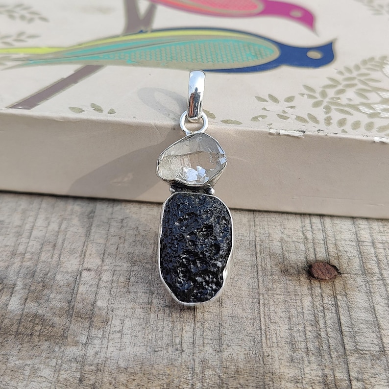 Puede incluir: Un colgante de plata con un cristal de cuarzo transparente y una piedra de obsidiana negra. El colgante est&aacute; sobre una superficie de madera.