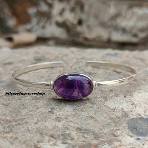 K&ouml;nnte beinhalten: Ein silbernes Armband mit einem zentralen, oval geformten violetten Amethyst-Edelstein. Das polierte Metallband hat ein offenes Design, und der Text silverringstoreshop ist in kleinen schwarzen Buchstaben unten links im Bild aufgedruckt.