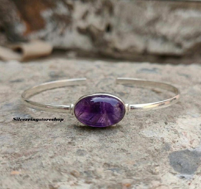 K&ouml;nnte beinhalten: Ein silbernes Armband mit einem zentralen, oval geformten violetten Amethyst-Edelstein. Das polierte Metallband hat ein offenes Design, und der Text silverringstoreshop ist in kleinen schwarzen Buchstaben unten links im Bild aufgedruckt.