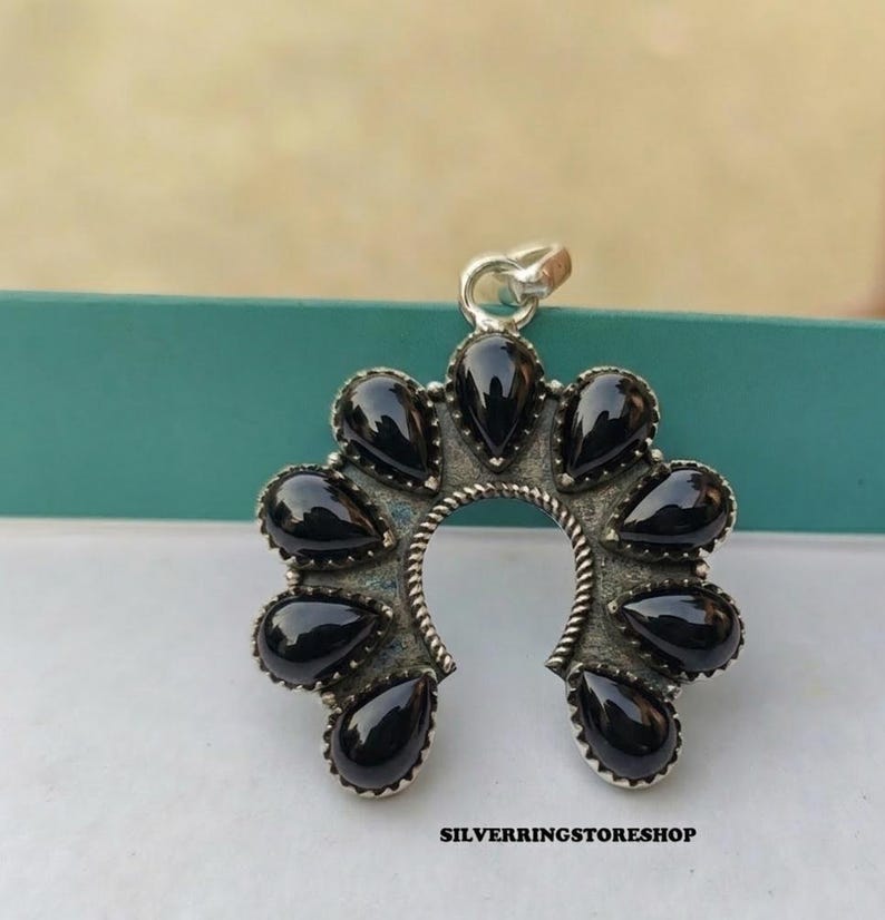 Pu&ograve; includere: Ciondolo in argento a forma di ferro di cavallo, ornato con pietre nere a forma di goccia. Il ciondolo ha una finitura strutturata e ossidata e un piccolo anello per una catena. Il testo "SILVERRINGSTORESHOP" &egrave; visibile in basso.