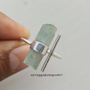 Könnte beinhalten: Ein silberner Ring mit einem hellblauen Aquamarin-Stein, der in einer silbernen Schiene gefasst ist. Die Schiene ist eine einfache, gerade Linie mit einem kleinen, senkrechten Balken, der von der Seite absteht.