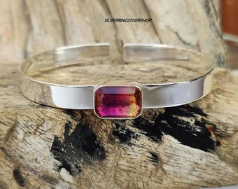 May include: A silver bangle bracelet with a pink and orange rectangular gemstone set in the center. The bracelet is on a wooden surface.
