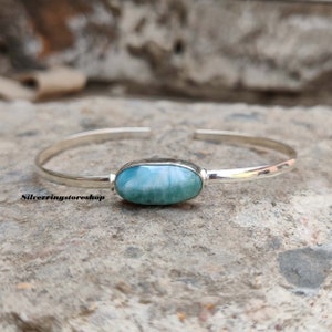 K&ouml;nnte beinhalten: Ein silbernes Armband mit einem zentralen, ovalen, blau-wei&szlig;en Larimar-Edelstein. Das polierte Metallband ist auf der R&uuml;ckseite offen. Der Text "Silverringstoreshop" ist in Schwarz auf der linken Seite des Armbands aufgedruckt.