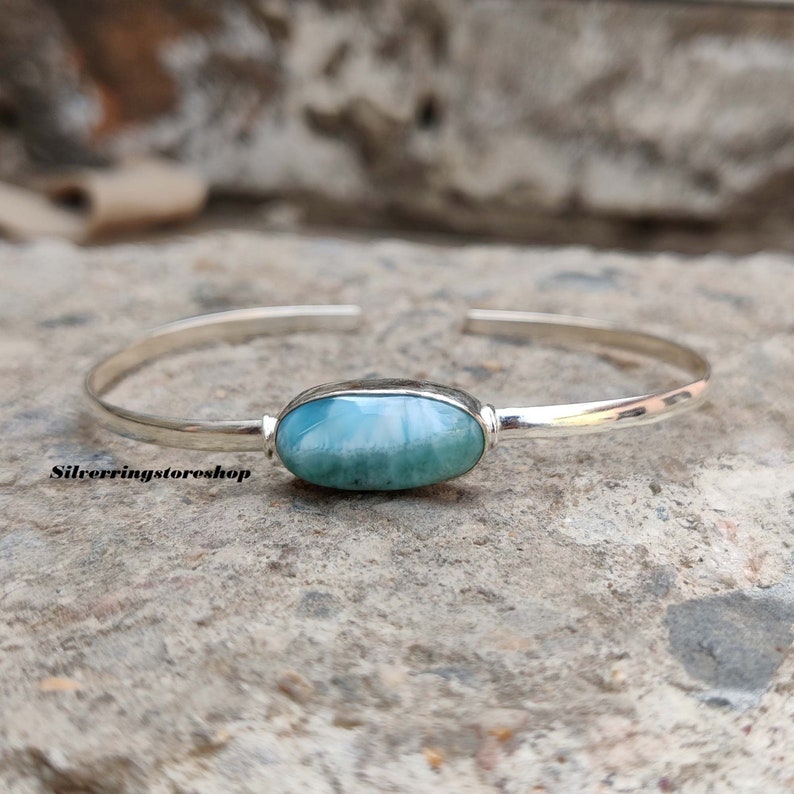 K&ouml;nnte beinhalten: Ein silbernes Armband mit einem zentralen, ovalen, blau-wei&szlig;en Larimar-Edelstein. Das polierte Metallband ist auf der R&uuml;ckseite offen. Der Text "Silverringstoreshop" ist in Schwarz auf der linken Seite des Armbands aufgedruckt.