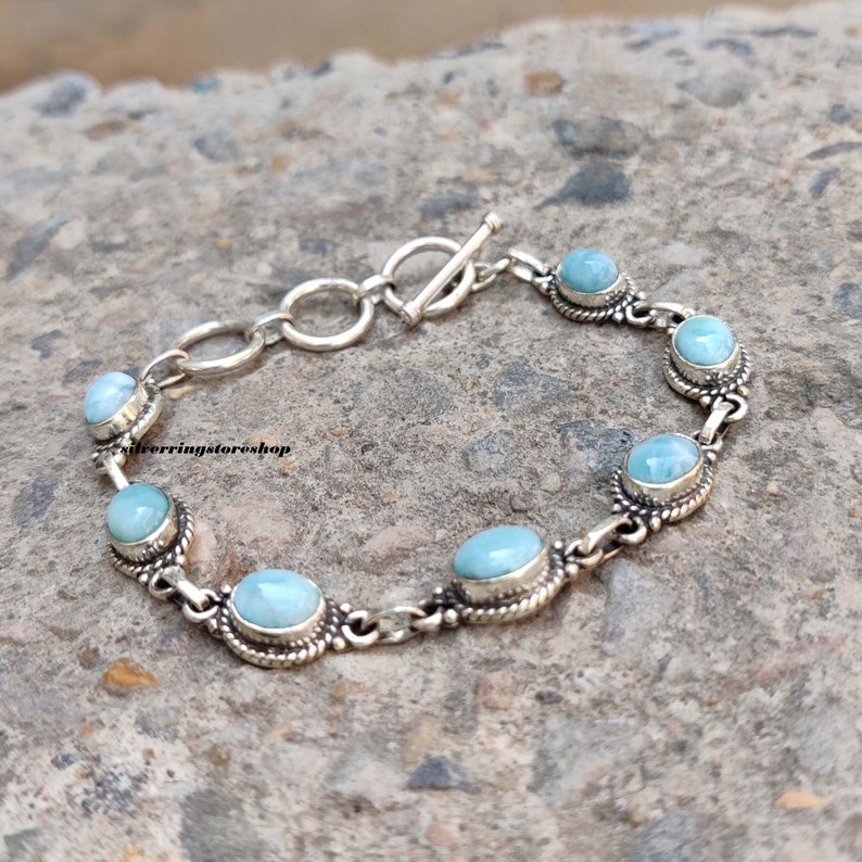 May include: A silver bracelet featuring oval, light blue gemstones. The bracelet has a toggle clasp and is composed of interconnected links. The gemstones are set in ornate, silver bezels, creating a delicate and elegant design.