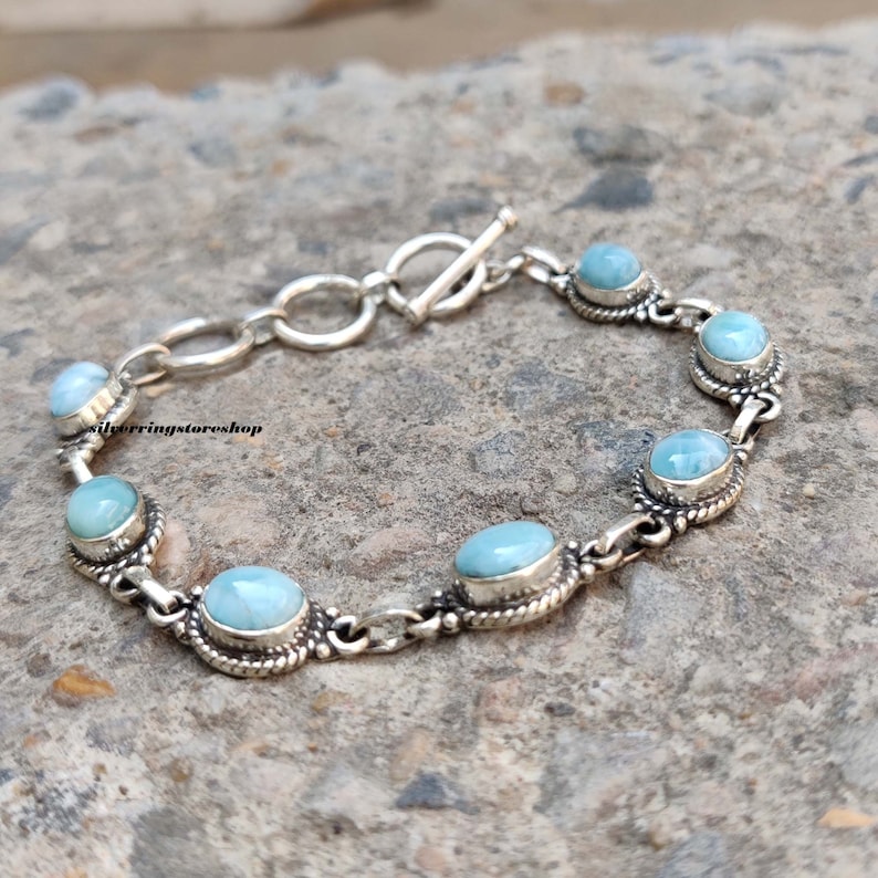 May include: A silver bracelet featuring oval and round light blue gemstones. The gemstones are set in ornate silver bezels and linked by a chain. The bracelet has a toggle clasp. The text "silverringstore shop" is visible.