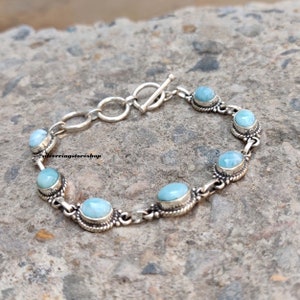 May include: A silver bracelet featuring oval, light blue gemstones. The bracelet has a toggle clasp and is composed of interconnected links. The gemstones are set in ornate, silver bezels, creating a delicate and elegant design.