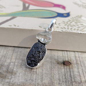 Puede incluir: Un colgante de plata con un cristal de cuarzo transparente sobre una roca volc&aacute;nica negra. El colgante est&aacute; sobre una superficie de madera.