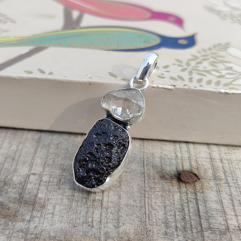 Puede incluir: Un colgante de plata con un cristal de cuarzo transparente sobre una roca volc&aacute;nica negra. El colgante est&aacute; sobre una superficie de madera.