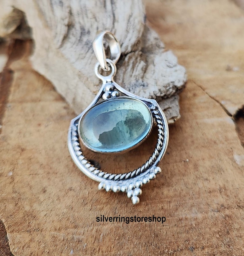 May include: A silver pendant with a light blue oval gemstone set in a decorative silver frame. The pendant has a delicate design with small beads around the edge.