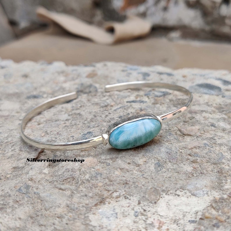 K&ouml;nnte beinhalten: Ein silberner Armreif mit einem einzelnen ovalen, hellblau und wei&szlig; marmorierten Larimar-Edelstein, der in einer glatten Metallfassung sitzt. Der Armreif hat eine polierte Oberfl&auml;che und ein offenes Design. Der Text silverringstoreshop ist sichtbar.