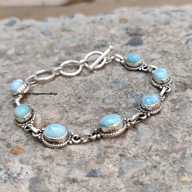 May include: A silver bracelet featuring oval and round light blue gemstones. The gemstones are set in ornate silver bezels and linked by a chain. The bracelet has a toggle clasp. The text "silverringstore shop" is visible.
