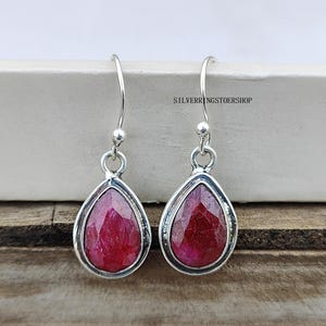 May include: Silver teardrop earrings featuring faceted, deep red gemstones. The earrings have a simple, elegant design with a polished finish. The text "SILVERRINGSTOERSHOP" is visible in the background.