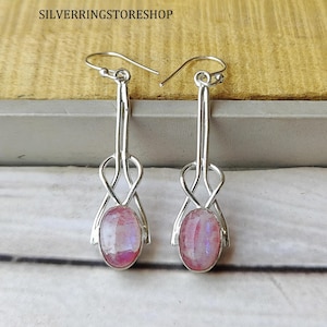 Pode incluir: Um par de brincos pendentes de prata com cabochões de pedra da lua ovais e iridescentes fixados na base de uma estrutura metálica fina e em laço. Os brincos estão suspensos por ganchos simples e apresentam um acabamento polido e simétrico.