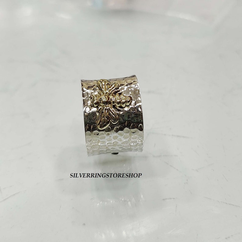 May include: A silver ring with a hammered texture and a gold bee design. The ring is wide and has a rounded shape.