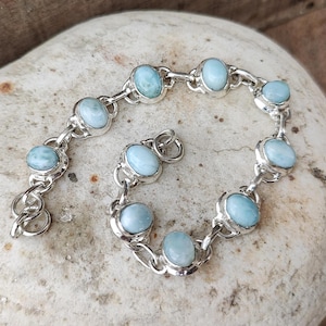 May include: A silver bracelet featuring oval, light blue gemstones. The gemstones are set in individual bezels and connected by silver links. The bracelet is displayed on a light-colored, textured surface.
