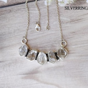 Op de afbeelding: Een zilveren ketting met een rij van zes heldere, gefacetteerde, ruw geslepen stenen. De ketting heeft een zilveren ketting en een karabijn sluiting.