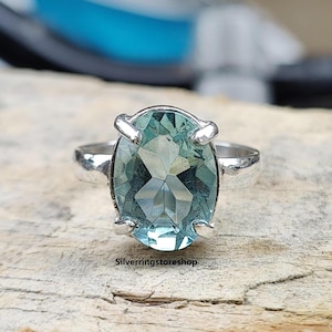 以下が含まれることがあります： 木製の表面に置かれた、薄い青色の楕円形の宝石がついた銀の指輪です。