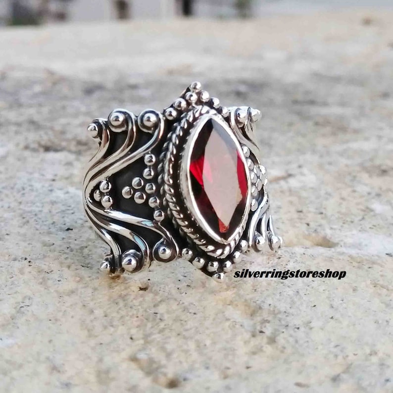 Puede incluir: Un anillo de plata con un dise&ntilde;o ornamentado que presenta una gran piedra preciosa roja. El anillo est&aacute; engastado con una piedra preciosa roja oscura de forma marquesa facetada. La banda est&aacute; decorada con detalles intrincados, que incluyen espirales, puntos y un patr&oacute;n de cuerda.