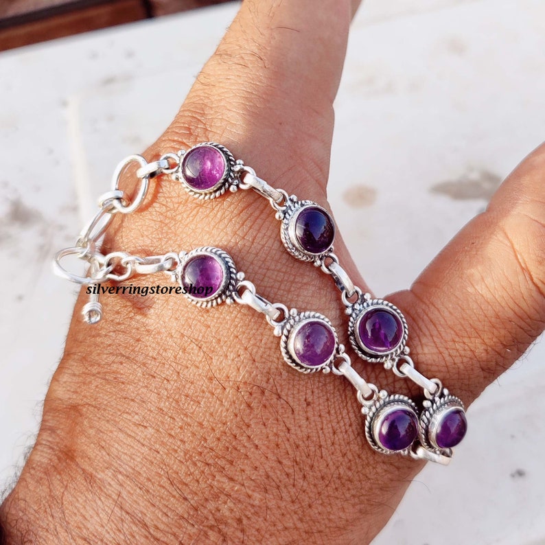 May include: A silver bracelet featuring multiple round, deep purple amethyst stones. The stones are set in ornate, circular bezels and connected by silver links. The bracelet is shown on a person's wrist.