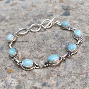 May include: A silver bracelet featuring oval and round light blue gemstones. The bracelet has a toggle clasp and is linked with small silver connectors. The gemstones are set in a decorative silver setting. The bracelet is on a gray surface.