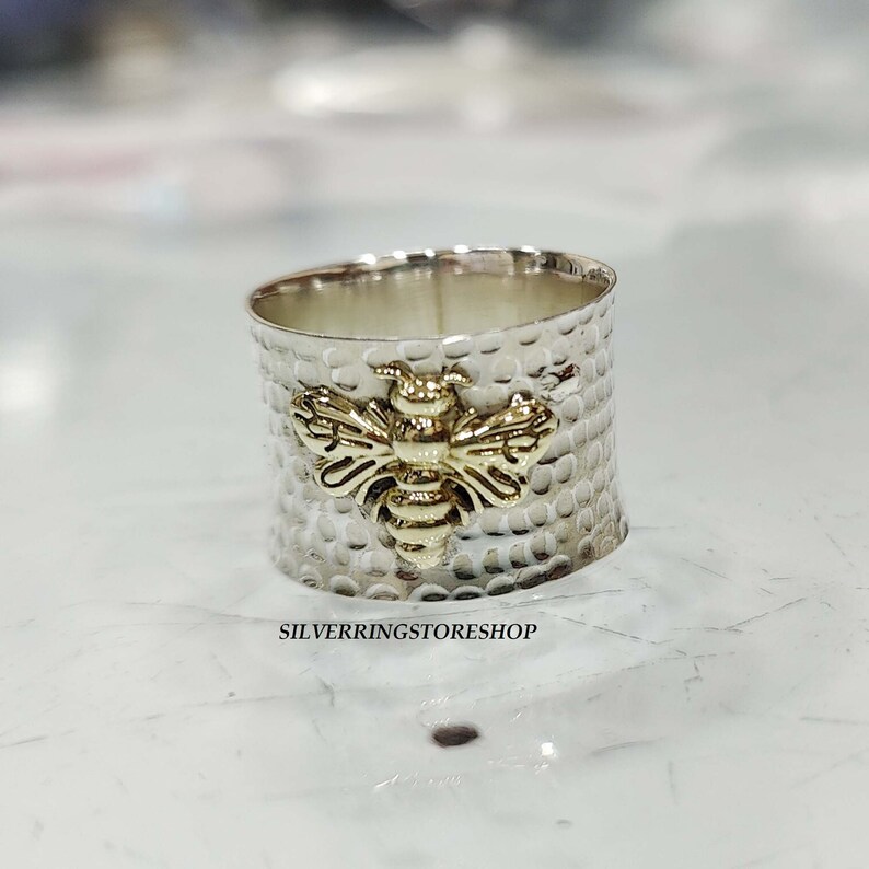 May include: A silver ring with a hammered texture and a gold bee design. The ring is wide and has a rounded shape.