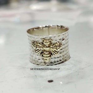 May include: A silver ring with a hammered texture and a gold bee design. The ring is wide and has a rounded shape.