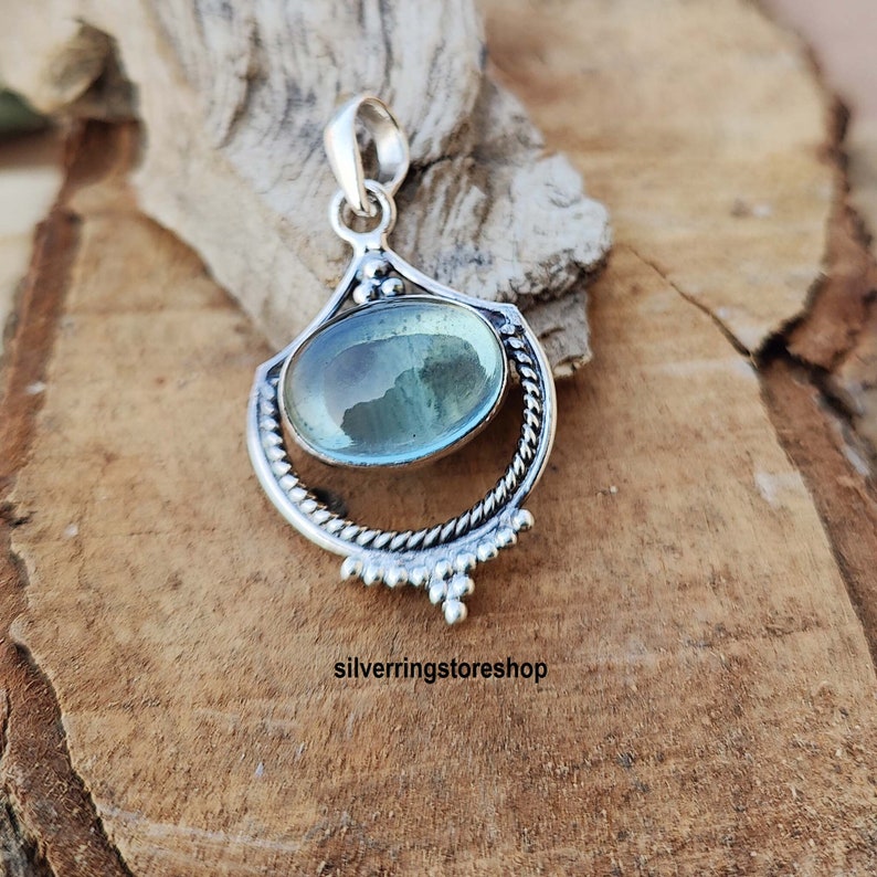 May include: A silver pendant with a light blue oval gemstone set in a decorative silver frame. The pendant has a bail for attaching to a chain.