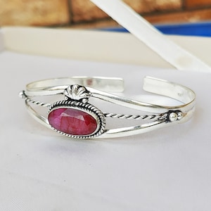 Puede incluir: Un brazalete de plata con una piedra ovalada rosa engastada en un engaste de plata. El brazalete tiene un diseño de alambre trenzado y un acento decorativo de plata.
