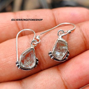 Pode incluir: Um par de brincos de prata com gotas de pedras preciosas transparentes, facetadas e cortadas em bruto. Os brincos têm um design simples e minimalista.