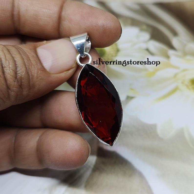 May include: A silver pendant with a large, deep red, faceted gemstone. The pendant is in the shape of a marquise.