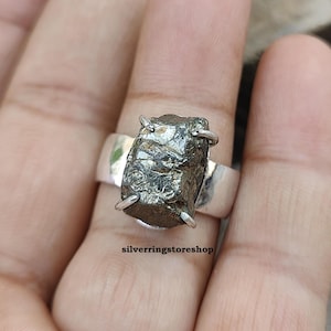 May include: A silver ring with a large, dark gray, rough-cut gemstone. The ring is set in a simple band and the gemstone is held in place by four prongs. The ring is on a finger.