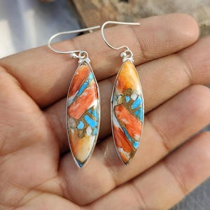 Pode incluir: Um par de brincos pendentes com um gancho de prata e uma pedra em forma de lágrima. A pedra é uma mistura de cores laranja, azul e branco.