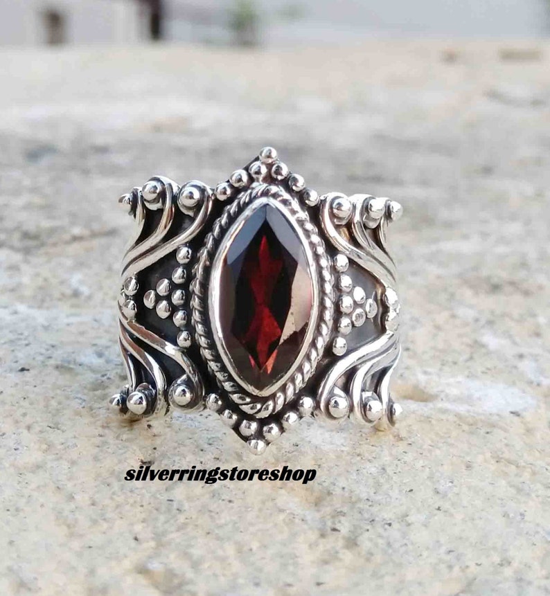 Puede incluir: Un anillo de plata con un dise&ntilde;o ornamentado que presenta una gran piedra preciosa de color rojo oscuro. El anillo tiene un dise&ntilde;o detallado e intrincado con peque&ntilde;as cuentas y remolinos.