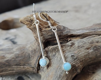 May include: A pair of silver dangle earrings with light blue gemstone accents. The earrings feature a long, thin silver bar with a round gemstone at the bottom.