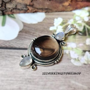 May include: A silver pendant with a large, round, smoky quartz stone at the center. Two smaller, oval-shaped moonstone accents flank the main stone. The pendant has a decorative, ornate design and the text "SILVERRINGSTORESHOP" is visible.