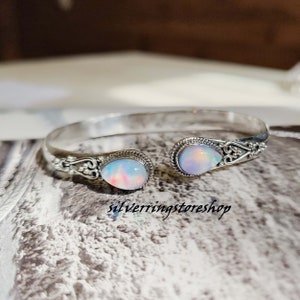 Puede incluir: Un brazalete de plata con dos piedras de ópalo iridiscente. El brazalete está abierto y tiene un diseño delicado.