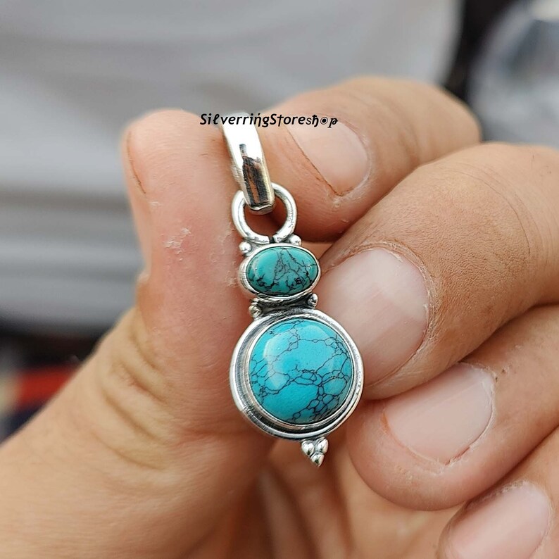 May include: A silver pendant with two turquoise stones. The larger stone is round and the smaller stone is oval. The pendant has a bail at the top for attaching to a chain.