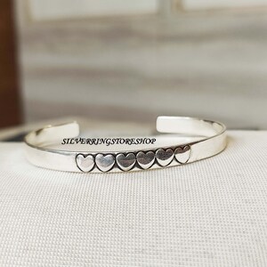 Könnte beinhalten: Ein silbernes Armreif-Armband mit einer Reihe herzförmiger Charms. Das Armband ist offen und hat ein schlichtes Design.