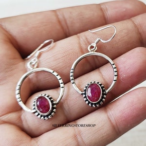 May include: A pair of silver hoop earrings with a red gemstone in the center of each hoop. The hoops are decorated with a beaded edge.